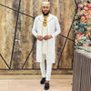 Man in a white traditional outfit with gold accents standing in front of a decorative wall with flowers.